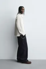 Camisa de corte relajado confeccionada en tejido de algodón, con diseño de rayas verticales. Presenta cuello solapa, cierre frontal de botones, manga larga con puños abotonados y un bolsillo plastrón en el pecho con un pequeño bordado decorativo.