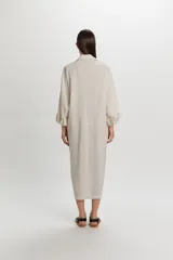 Vestido camisero midi color beige con sutiles rayas verticales oscuras, de manga larga, cuello camisero, cierre frontal con botones y detalle de fruncidos drapeados en los laterales de la falda.