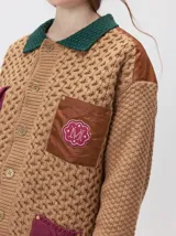 Campera tejido grueso y abrigado color verde militar, con forro de nylon interno, cuello estilo camisa en color negro y cierre frontal con botones. Cuenta con cuatro bolsillos delanteros con apliques en distintos tonos y texturas, y un parche bordado circular en el bolsillo superior izquierdo.