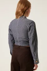 Camisa corta a rayas verticales grises y blancas, con cuello clásico y mangas largas. Presenta un diseño trenzado en el frente con botones dorados y aberturas.