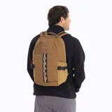 Mochila Merrell modelo Trout Basic de 25 litros, color marrón. Presenta un diseño funcional con compartimento principal con cierre, bolsillos laterales elásticos con correas de ajuste y una cinta frontal decorativa. Confeccionada en poliéster resistente con recubrimiento de PU.