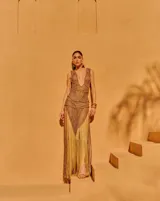 Vestido largo de tejido crochet en color bronce, con escote en V profundo, diseño de red y detalles trenzados. Presenta una silueta ajustada y un acabado con flecos largos en el ruedo.