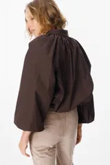 Camisa de corte holgado color marrón chocolate, con cuello clásico, cierre frontal de botones y mangas largas abullonadas con frunces en los hombros.