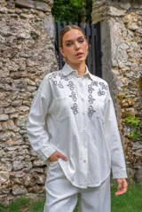Camisa blanca de algodón con cuello clásico, cierre frontal con botones metálicos a presión, dos bolsillos de parche en el pecho y apliques de brillos en la parte delantera.