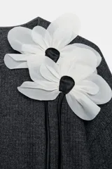 Chaqueta gris de punto con cuello redondo, manga larga y cierre frontal con botones. Detalle de flor blanca y negra en el frente. Tejido en rib.