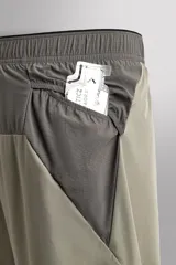 Pantalón corto de trail running color beige, confeccionado en tejidos técnicos ligeros y elásticos. Cuenta con forro interior, cintura elástica ajustable con cordón, bolsillos traseros laterales multifunción, bolsillo trasero central con cremallera, bajo con costura termofijada y aberturas laterales. Incluye logo estampado en la pernera.