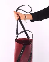 Matera tipo bucket de cuero color bordó, con detalles de tiras verticales en cuero negro adornadas con remaches metálicos plateados. Cuenta con un asa de mano fija y un compartimento interior amplio.