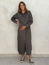 Vestido camisero largo color gris oscuro, de manga larga con presilla para sujetar el puño, cuello camisero y cierre frontal de botones. Presenta bolsillos de parche en el pecho y aberturas laterales en el ruedo.