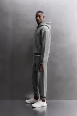 Sudadera hoodie gris de corte relaxed fit, confeccionada en tejido de algodón con interior afelpado. Presenta cuello con capucha ajustable, manga larga, texto estampado en color bordó en el pecho, bolsillo frontal tipo canguro, aplicación de parche con logo en la parte inferior y acabados en rib.
