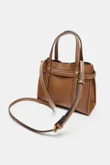 Bolso tipo mini shopper de cuero sintético color marrón caramelo, con costuras blancas visibles. Posee asas de mano y una correa bandolera ancha de tela color beige. El frente tiene un detalle de tira con hebilla metálica dorada.