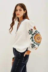 Camisa blanca de doble gasa de algodón, corte amplio y mangas largas abullonadas. Presenta bordados a mano en mangas y escote con motivos florales en verde, amarillo y violeta.
