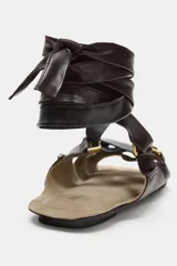 Sandalia plana de cuero color marrón oscuro, con tiras cruzadas decoradas con apliques metálicos dorados y cierre mediante tiras largas para atar al tobillo.
