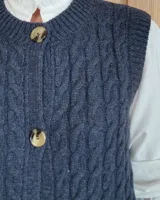 Chaleco de punto con diseño de trenzas, color gris oscuro, con cierre frontal de botones grandes y bolsillos laterales.