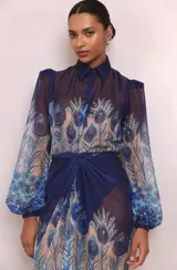 Camisa de organza semitransparente con estampado de plumas de pavo real en tonos azules y cuello doble. Cuenta con mangas abullonadas con abertura y puños cerrados con botón. Incluye hombreras.