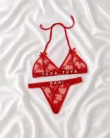 Conjunto de lencería rojo confeccionado en tul bordado, compuesto por un soutien triangular con tirantes finos para anudar al cuello, moños decorativos y aplicaciones de letras brillantes con la frase "TODA TUYA", y una bombacha colaless con la palabra "DANI".