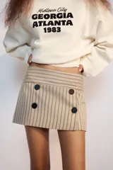 Falda pantalón de tiro bajo color beige con diseño de rayas diplomáticas verticales en tonos oscuros, con detalles de cuatro botones negros decorativos en la parte frontal y cierre lateral con cremallera oculta.