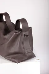 Cartera de cuero color marrón, con diseño suave y flexible. Cuenta con cierre metálico superior, doble asa corta para llevar al hombro y una correa larga regulable y desmontable. Interior forrado con bolsillos organizadores.