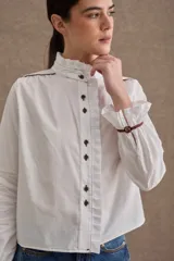 Camisa de popelina blanca con cuello alto tipo victoriano, volados frontales y detalles de costuras y ribetes en color marrón. Presenta mangas largas con puños elásticos terminados en volado y cierre frontal con botones oscuros.