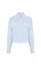 Camisa de corte cropped y entallado, color celeste, con cuello clásico, mangas largas con puños, cierre frontal de botones y dos bolsillos delanteros con solapa y botón.