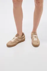 Zapatillas urbanas de corte bajo en tonos beige y tostado, combinando paneles de cuerina y gamuza sintética. Cuentan con cordones anchos tipo cinta de raso en color crudo, pespuntes decorativos y suela de goma al tono.
