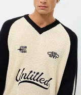 Suéter de punto con corte boxy, diseño bicolor en crema y negro. Presenta cuello en V, mangas largas con rayas laterales y estampa frontal con lettering Untitled y detalles gráficos. Cuenta con terminaciones acanaladas a rayas en el ruedo y puños.