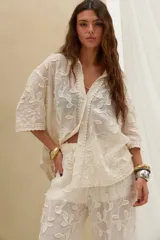 Camisa de manga corta color beige con bordado floral en relieve, cierre frontal con botones y detalles de encaje en los bordes.