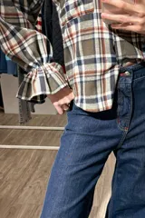 Camisa de manga larga a cuadros en tonos verde oliva, blanco y naranja. Presenta cuello camisero, cierre frontal con botones, mangas abullonadas y puños elásticos con detalle de volado.