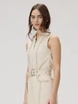 Enterito sin mangas confeccionado en denim de algodón color beige, de fit holgado con pierna recta y tronco entallado. Posee cierre frontal completo, cuello camisero y detalles de tachas metálicas en las costuras. Incluye un cinturón ajustable del mismo material con hebilla rectangular.
