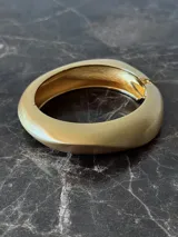 Pulsera rígida dorada con forma ovalada irregular y cierre a presión.