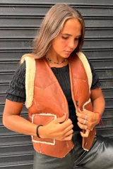 Chaleco de cuero genuino color tostado con interior y detalles en piel sintética color crema. Presenta cierre frontal con botones, dos bolsillos delanteros y diseño con costuras visibles.