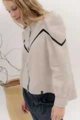 Camisa de lino color beige con mangas abullonadas y detalles de puntilla negra en forma de V en el frente. Presenta cuello a la base con borde desflecado, cierre frontal con botones forrados y puños con alforzas.