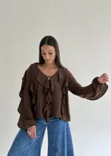 Blusa blanca de corte holgado con escote en V, decorada con volados frontales y en las mangas. Presenta mangas largas con puños elásticos y cierre frontal con botones.