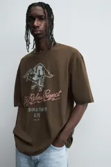 Camiseta de algodón color marrón oscuro, corte relajado (relaxed fit) y manga corta. Presenta un estampado frontal con un lobo o cánido corriendo, el texto "The Riders Project", "VISION & TEMPER" y "0.79 per gallon" en tonos grises y rosados.