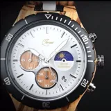 Reloj cronógrafo con caja de madera de zebrano y bisel técnico de acero negro. La esfera es plateada con subdiales de madera, indicador de fase lunar y fechador. La malla combina eslabones de acero inoxidable con madera de zebrano.