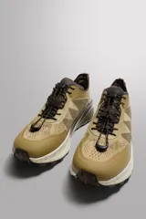 Zapatilla deportiva de trail running color beige con detalles en marrón. Presenta una parte superior de tejido técnico transpirable, refuerzos en puntera y talón, sistema de ajuste rápido con cordones técnicos y entresuela con tecnología de amortiguación y acabado moteado.