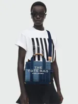 Cartera tipo tote de Marc Jacobs, confeccionada en denim con diseño de paneles en diferentes tonos de azul. Presenta el logo de la marca estampado en el frente, asas superiores de mano y una correa de hombro ajustable y extraíble en color azul oscuro.