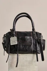 Cartera de cuero de terminación gastada y cuerpo blando, con dos asas de mano y correa desmontable para llevar cruzada. Cuenta con bolsillos exteriores con cierre, tiras colgantes y detalles de hebillas. Incluye un llavero metálico decorativo con la inscripción "WE THE FREE".