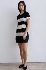 Vestido corto de corte recto con diseño a rayas horizontales en blanco y negro. Presenta franjas plisadas que aportan textura, escote redondo con detalle fruncido y mangas cortas con volados.