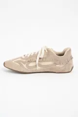 Zapatilla de cuero legítimo en tonos beige y crema, con diseño de flecos en el lateral y costuras decorativas visibles en el contorno. Presenta cordones al tono y suela plana.