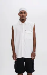 Camisa sin mangas de corte oversize, color blanco, con cuello clásico, cierre frontal con botones ocultos y bolsillo frontal con solapa y remaches metálicos.