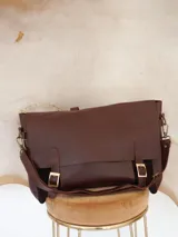 Maletín tipo cartero con solapa de cuero marrón y cuerpo de gamuza azul. Cuenta con correas ajustables con hebillas metálicas, asa superior y correa larga para llevar al hombro.