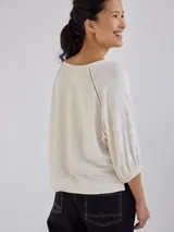 Remera de corte holgado en color crudo con sutiles rayas horizontales. Presenta mangas tres cuartos con puños elásticos, cuello redondo con detalle de costura en V y costuras decorativas oscuras en los hombros.