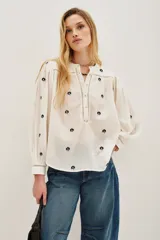 Camisa de algodón color crudo con bordados florales pequeños en azul marino. Presenta mangas largas abullonadas, cuello redondo y una abertura frontal con botones y detalles de encaje calado en la pechera y hombros.