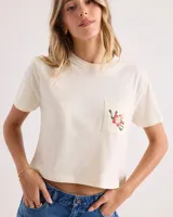 Remera corta de corte boxy en color blanco vintage, con mangas cortas, cuello redondo y un bolsillo aplicado en el pecho que cuenta con un pequeño estampado floral y de serpiente.