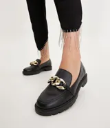Mocasines negros de cuero sintético con suela track y detalle de cadena dorada.