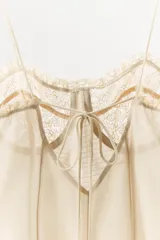 Vestido mini beige con aros de escote redondo y tirantes finos. Detalle de tejido combinado y costuras marcadas, con aplicación de encaje a tono y cierre en espalda con lazada.