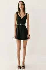 Vestido corto negro sin mangas, con escote en pico profundo y falda con pliegues. Incluye un cinturón fino de cuero sintético con hebilla metálica dorada en la cintura. Cierre lateral con cremallera oculta.