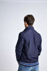 Campera liviana de nylon color gris oscuro, con cierre frontal completo, cuello subido y puños y cintura acanalados con rayas en rojo y blanco. Presenta logo bordado en el pecho y bolsillos laterales con cierre.