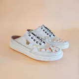 Calzado destalonado de cuero en color natural, con cordones y detalle de pintura artesanal en la puntera. Incorpora suela de goma blanca y aplique metálico lateral con forma triangular.