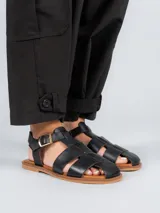 Sandalias planas de cuero negro estilo pescador, con tiras anchas entrelazadas en el empeine, suela marrón y tira ajustable con hebilla metálica en el tobillo.